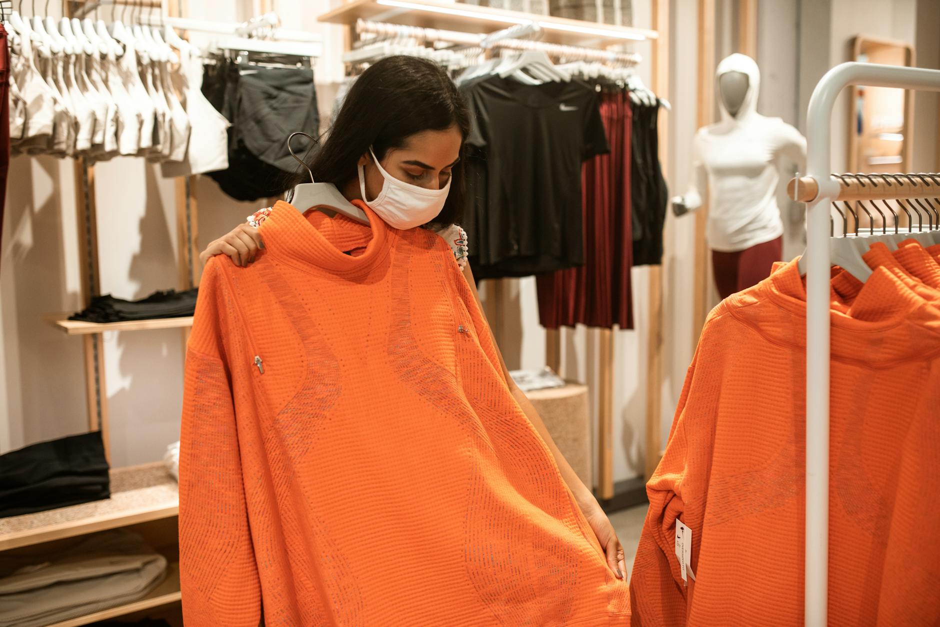 Kobieta w maseczce mierzy pomarańczową kurtkę w sklepie odzieżowym