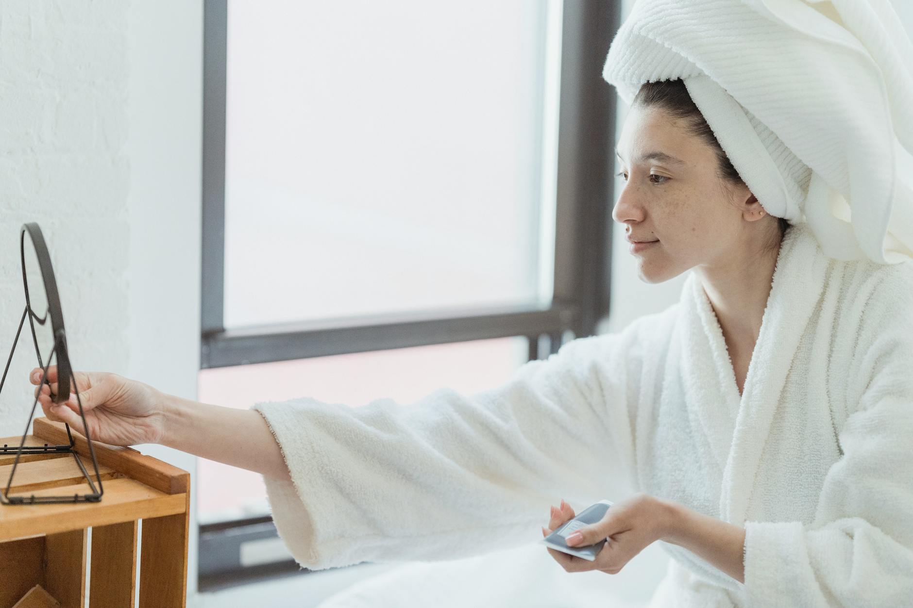 Kobieta w szlafroku nakłada kosmetyk na twarz przed lustrem