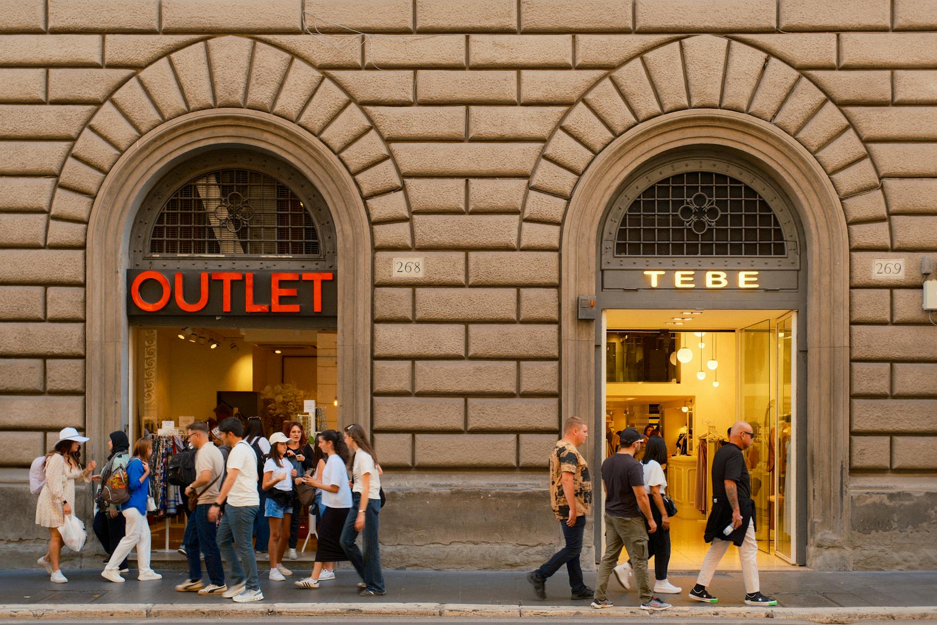 Czy warto kupować „outlet” na platformach ogłoszeniowych? Przechodnie przed sklepami outletowymi we Florencji pod arkadami