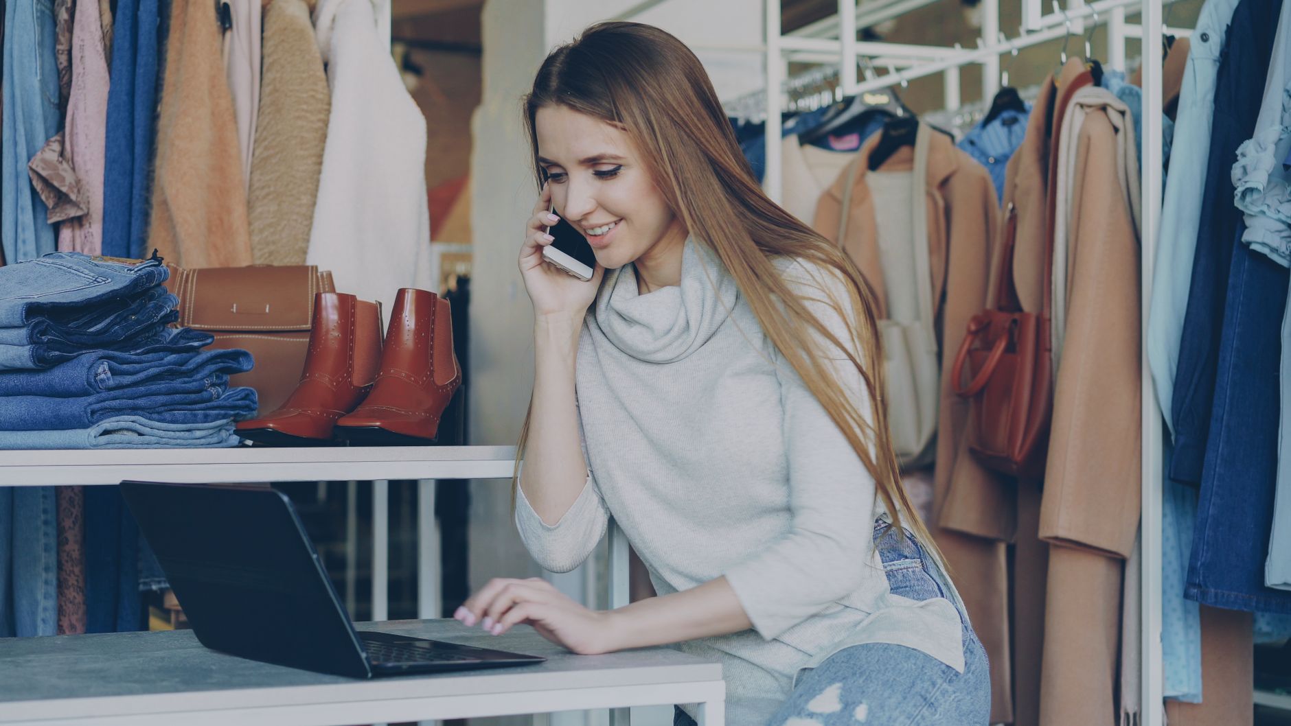 Uśmiechnięta kobieta w butiku odzieżowym korzysta z laptopa i telefonu